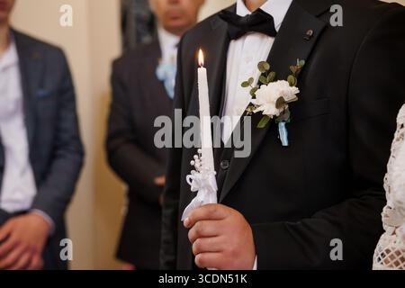 Eine Nahaufnahme eines Bräutigams in einem schwarzen Smoking, der während einer Hochzeitszeremonie eine Kerze in der Hand hält, umgeben von elegant gekleideten Gästen Stockfoto