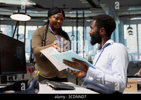 Manager im Büro übergibt Papiere an Mitarbeiter, die mit KI-Assistenten am PC arbeiten. Mitarbeiter, der von Teamleitern überwacht wird, während künstliche Intelligenz verwendet wird, um administrative Aufgaben zu optimieren, Kamera A. Stockfoto