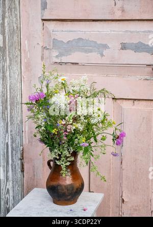Rustikaler Blumenstrauß aus wilden Wiesenblumen, einschließlich Kornblumen, Lupinen und rosa Astern, angeordnet in einem alten Tonkrug auf einem verwitterten Holztisch Agave Stockfoto