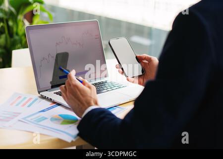 Unwiedererkennbarer eleganter junger Geschäftsmann in einem Anzug, der in einem Smartphone mit weißem Modell arbeitet, leerer Bildschirm und Laptop in einem Café in der Nähe Stockfoto