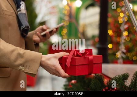 Mans Hand hält rote Geschenkbox als Geschenk verschönert und mit Smartphone. Weihnachtsfee. Weihnachtseinkäufe. Seitenansicht. Nahaufnahme. Stockfoto