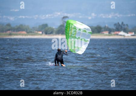 Ein Mann mittleren Alters in einem Neoprenanzug steht im Wasser, hält einen großen grünen und weißen Drachen mit einer Küstenlinie in der Ferne Stockfoto