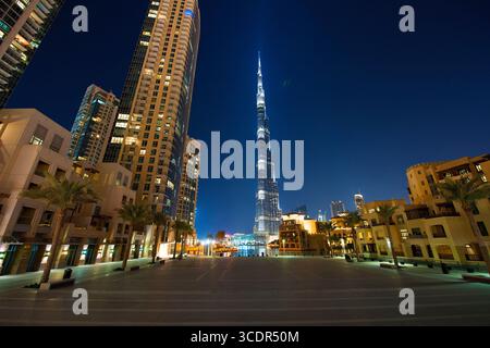 Moderne Stadtlandschaft der Innenstadt von Dubai bei Nacht mit beleuchtetem Burj Khalifa, umgeben von hohen Türmen, Palmen und urbaner Architektur. Stockfoto