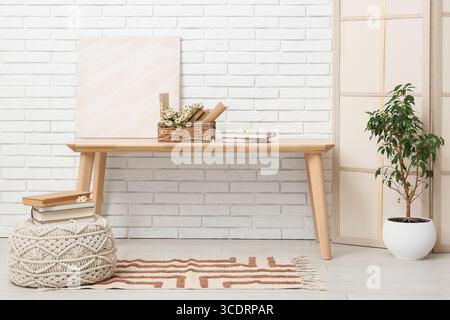 Korb mit Büchern, Kamillen und Gemälden auf dem Tisch in der Nähe der weißen Ziegelwand im Zimmer Stockfoto