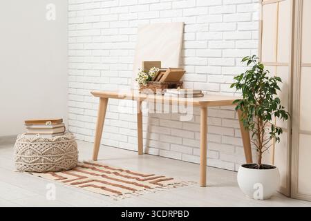 Korb mit Büchern, Kamillen und Gemälden auf dem Tisch in der Nähe der weißen Ziegelwand im Zimmer Stockfoto