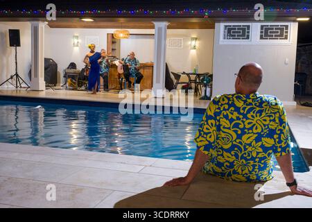 Mann, der am Rand des Schwimmbades sitzt und einen Jungen beobachtet Frau trägt ein blaues Kleid und tanzt zu einem hawaiianischen Hula Live-Musik von zwei reifen männlichen m Stockfoto