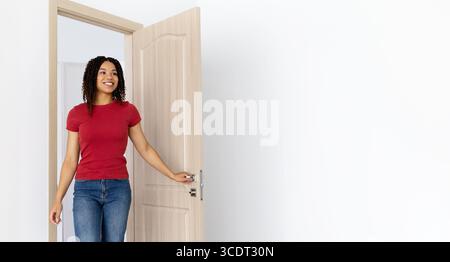 Lächelnde Afroamerikanerin in rotem T-Shirt und Jeans, die in ein helles Zuhause kommt Stockfoto