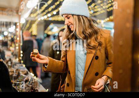 Glückliche junge Frau lächelt, während sie nachts eine kleine goldene Geschenkbox an einem festlichen Weihnachtsmarkt im Freien auswählt und hält. Stockfoto