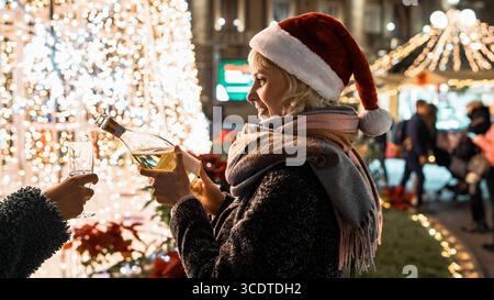 Glückliche junge Frau in einem Weihnachtsmann-Hut lächelt und spritzt Sekt in das Glas eines Freundes bei einer festlichen Weihnachts- oder Silvesterparty im Freien. Stockfoto