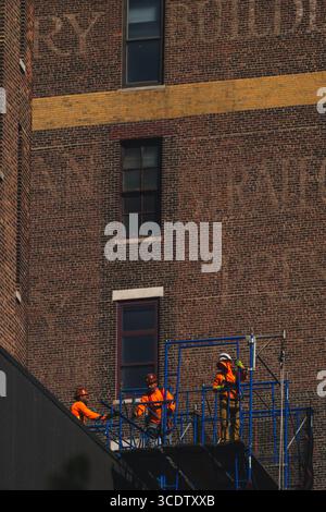 Blick auf Bauarbeiter in leuchtend orangefarbenem Getriebe auf einer blauen Gerüstplattform vor dem Hintergrund eines verwitterten Ziegelgebäudes in New York, New York, USA. Stockfoto