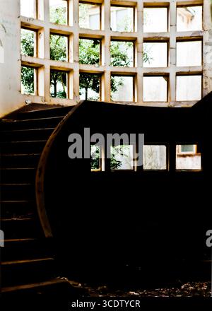 Eine Innenaufnahme eines Gebäudes mit einer Treppe und einem großen Fenster mit einem Gittermuster. Die Treppe ist dunkel und führt zum Fenster, das Fil ist Stockfoto
