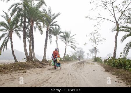 Jhenaidah, Bangladesch - 20. Januar 2024: Blick auf eine Mutter und ein Kind, die eine staubige Straße entlang laufen, gesäumt von schlanken Palmen, die Szene gedämpft von einem trüben Himmel. Stockfoto