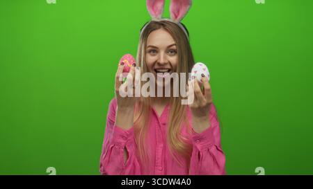 Junge Frau mit Hasenohren hält ostereier vor einem hellgrünen Hintergrund und lächelt fröhlich über farbenfrohe Dekorationen in festlicher Kleidung. Stockfoto