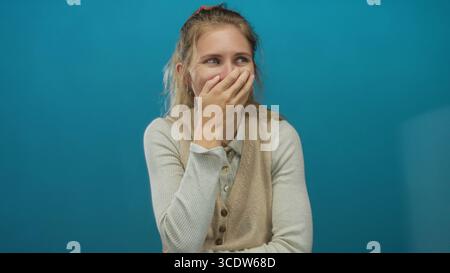 Die Frau, die die Hand benutzt, während sie auf einem Studio-Set lacht, zeigt einen blonden Stil und eine junge Atmosphäre, die einen blauen Hintergrund mit einer fröhlichen Lächelgeste bedeckt. Stockfoto