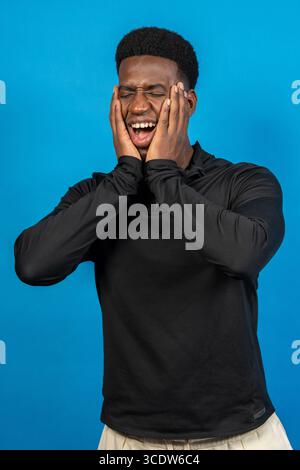 Aufgeregt, junger Mann, der sein Gesicht mit den Händen hält und vor Freude schreiend vor einem leuchtend blauen Hintergrund Stockfoto