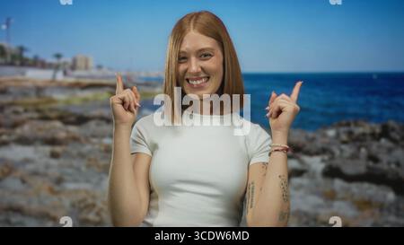 Die junge blonde Frau zeigt die Zeigefinger nach oben auf die felsige Küste an der sonnigen Küste; Optimismus Begeisterung Aufregung Freude. Stockfoto