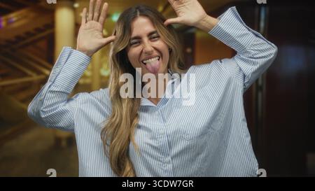 Die elegante blonde Frau spreizt die Arme mit einem hellen Lächeln auf und neigt den Kopf in einer mit Messingsäulen und warmen Deckenleuchten geschmückten Hotellobby. Playfu Stockfoto