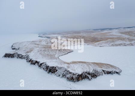 Aus der Vogelperspektive auf eine schneebedeckte Landschaft mit zerklüfteten Klippen, die auf einen gefrorenen See unter hellem Himmel treffen, den Baikalsee, Russland. Stockfoto