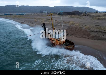 Aus der Vogelperspektive eines rostigen Schiffswracks, das an einem Sandstrand gestrandet ist, während Wellen um ihn herum krachen, mit Gebäuden im Hintergrund, Teriberka, Russland. Stockfoto