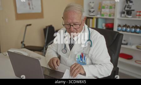 Senior Mann in einer Arztuniform arbeitet drinnen in einer Klinik, untersucht Dokumente mit einem Stethoskop sichtbar, repräsentiert eine professionelle Gesundheitsfürsorge e Stockfoto