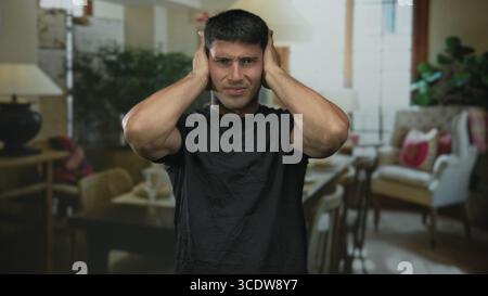 Der Mann bedeckt die Ohren mit beiden Händen zwischen gemütlichen Wohnzimmerstühlen und Lampe in einem Gebäudeinneren; Distress. Stockfoto