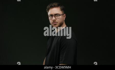 Ein junger Mann mit Bart und Brille steht selbstbewusst vor einer schwarzen Wand mit einem isolierten und konzentrierten Verhalten. Stockfoto