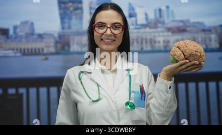 Frau Ärztin mit Hirnmodell an der Strandpromenade mit Stethoskop in Uniform im Freien, selbstbewusst lächelnd. Stockfoto