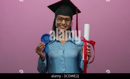 Lächelnde Frau in Abschlusskleidung mit Diplom und Band vor rosa Hintergrund feiert akademische Leistungen. Stockfoto