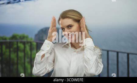 Frau lächelt draußen in der Natur mit Blick auf das Meer, trägt ein weißes Hemd, begrüßt fröhlichen, fröhlichen Ausdruck, fängt Glück und Freiheit ein Stockfoto