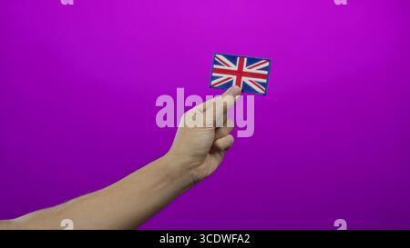 Die männliche Hand hält einen gestickten unions-Jack vor rosa Hintergrund, der die Identität großbritanniens und den kulturellen Stolz symbolisiert, isoliert mit lebhafter Betonung. Stockfoto