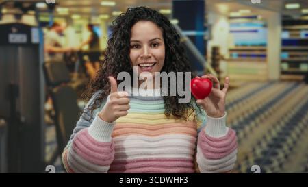 Die lächelnde Frau hält das Herz, während sie im Fitnessstudio mit einem farbenfrohen Pullover die Daumen nach oben drückt und im Fitnesscenter positiver wirkt. Stockfoto