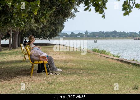 Grado, Italien - 24. Juni 2025: Ein friedlicher Moment auf Barbana Island: Eine Frau sitzt ruhig an der Lagune, während ein Mann auf einer gelben Bank ruht Stockfoto