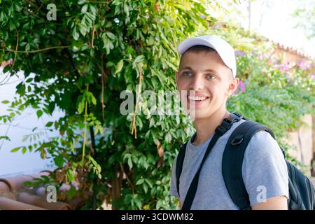 Ein junger Mann mit weißem Hut und grauem Hemd lächelt. Er hält einen Rucksack und steht vor einem Busch Stockfoto