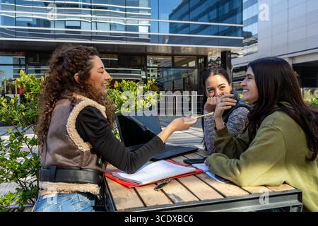 Drei glückliche Universitätsstudenten studieren und teilen sich Notizen an einem Tisch draußen und genießen ihre gemeinsame Zeit Stockfoto