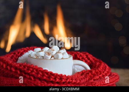 Tasse mit heißer Schokolade und Marshmallows in rotem Schal vor dem Kamin Stockfoto