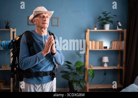 Meditation. Pensionierter Reisender mit Rucksack steht in meditativer Pose, genießt friedliche Hände in betenden Gesten im Innenraum. Touristen machen aktiv Stockfoto