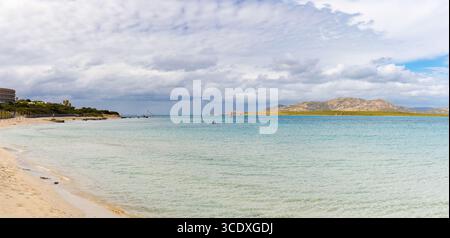 Panoramablick auf den Strand La Pelosa auf Sardinien, mit türkisfarbenem, flachem Wasser, Sandstrand und fernen felsigen Hügeln unter bewölktem Himmel. Stockfoto