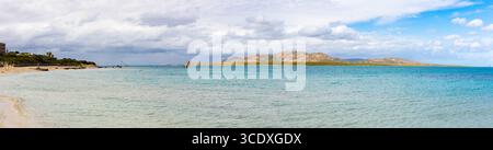 Großer Panoramablick auf den Strand La Pelosa auf Sardinien, mit türkisfarbenem, flachem Wasser, Sandstrand und felsigen Hügeln unter teilweise bewölktem Himmel. Stockfoto