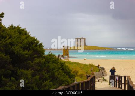 Blick auf den Torre della Pelosa vom Strand in Sardinien, mit türkisfarbenem Wasser, Sandstrand und bewölktem Himmel umgeben von Küstenvegetation. Stockfoto
