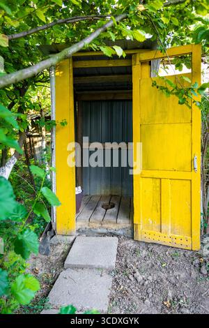 Rustikales gelbes hölzernes Outthouse unter grünen Bäumen mit offener Tür, Tageslicht und Holzboden in ländlicher Gartenumgebung. Stockfoto