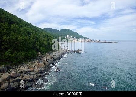 Ein bewaldeter Hügel fällt in eine felsige Küste, während sich die Küste in Richtung Cheongsapo Hafen biegt, wo die rot-weißen Leuchttürme am Breakwat stehen Stockfoto
