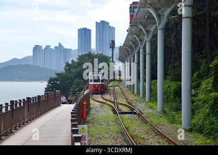 Zwei Kutschen der Haeundae Blueline Park Küstenbahn fahren an der Promenade nahe Songjeong vorbei, während die Himmelskapsel über und HAEU verläuft Stockfoto