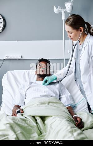 Kaukasische Frau mit Labormantel, die im modernen Krankenhaus arbeitet und den Gesundheitszustand des jungen Schwarzen Mannes mit Stethoskop überprüft Stockfoto