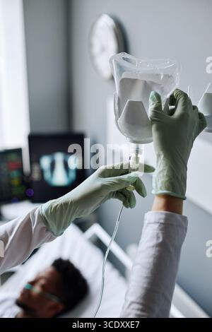 Mittlere Nahaufnahme einer nicht erkennbaren Krankenschwester mit Latexschutzhandschuhen, die den IV-Beutel für den Patienten auf der Krankenhausstation wechseln Stockfoto