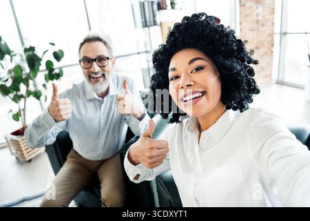 Zwei verschiedene Kollegen am Arbeitsplatz lächeln und zeigen die Daumen hoch, was Teamarbeit und positive Einstellung repräsentiert Stockfoto