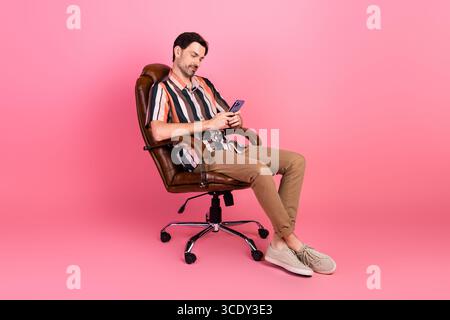 Entspannter Mann in einem stilvollen gestreiften Hemd, der auf einem ledernen Bürostuhl sitzt und ein Smartphone mit einem fröhlichen Ausdruck benutzt Stockfoto