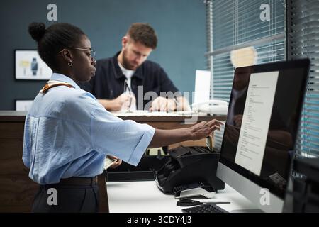 Schwarze Angestellte, die am Computerterminal im Bankbüro arbeitete, während ein kaukasischer Mann am Schalter stand und Papiere ausfüllte, beide konzentrierten sich auf den Finanztransaktionsprozess Stockfoto