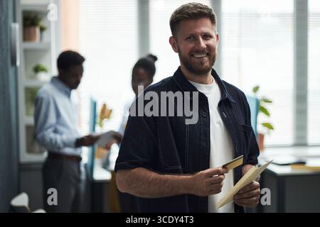Kaukasischer junger Mann lächelt, während er Kreditkarte und Dokument im Bankbüro hält, zwei schwarze junge männliche und weibliche Kollegen diskutieren Papierkram im Hintergrund, moderner Arbeitsbereich sichtbar Stockfoto