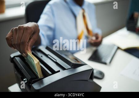 Hand des Bankarbeiters, der Bargeld mit einem Rechnungszählgerät im Büro verarbeitet, Banknoten von Hand einlegt, während er am Schreibtisch sitzt und die Computertastatur und Dokumente sichtbar ist Stockfoto