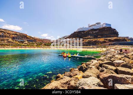 Felsige Atlantikküste mit Sandstrand, Touristen, bunten Tretbooten und RIU Hotel auf felsigen Klippen. Spanien. Grand Canaria. Stockfoto